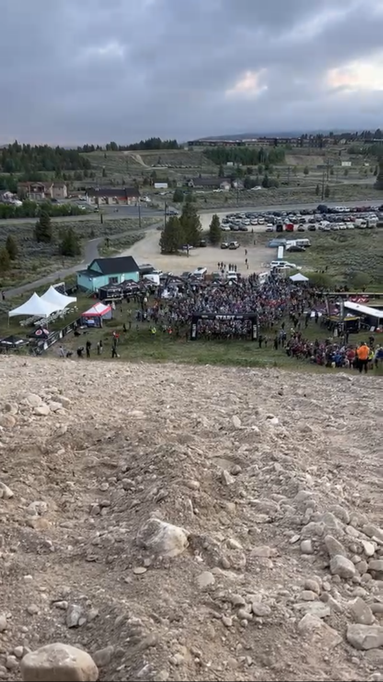 Large crowd gathered in a dirt field with buildings in the background at dusk.