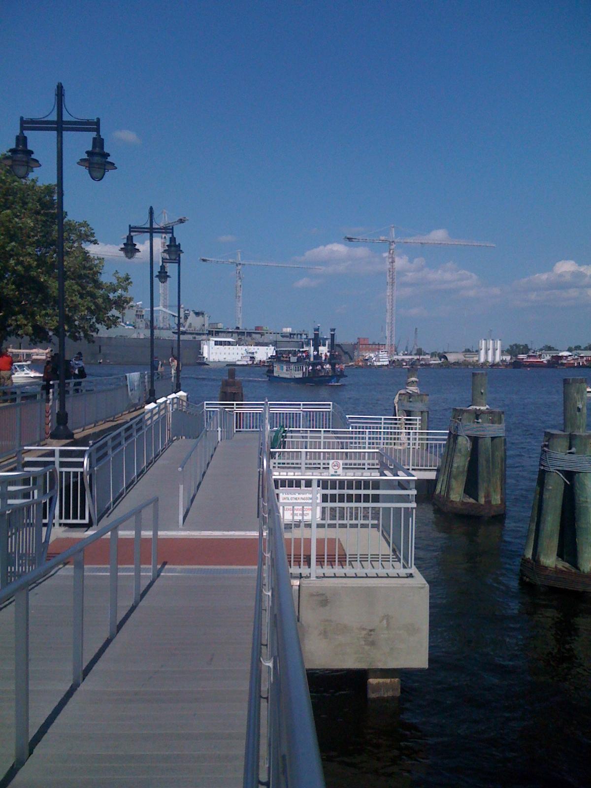 A wooden pier extends into a sunny bay with boats and distant cranes.