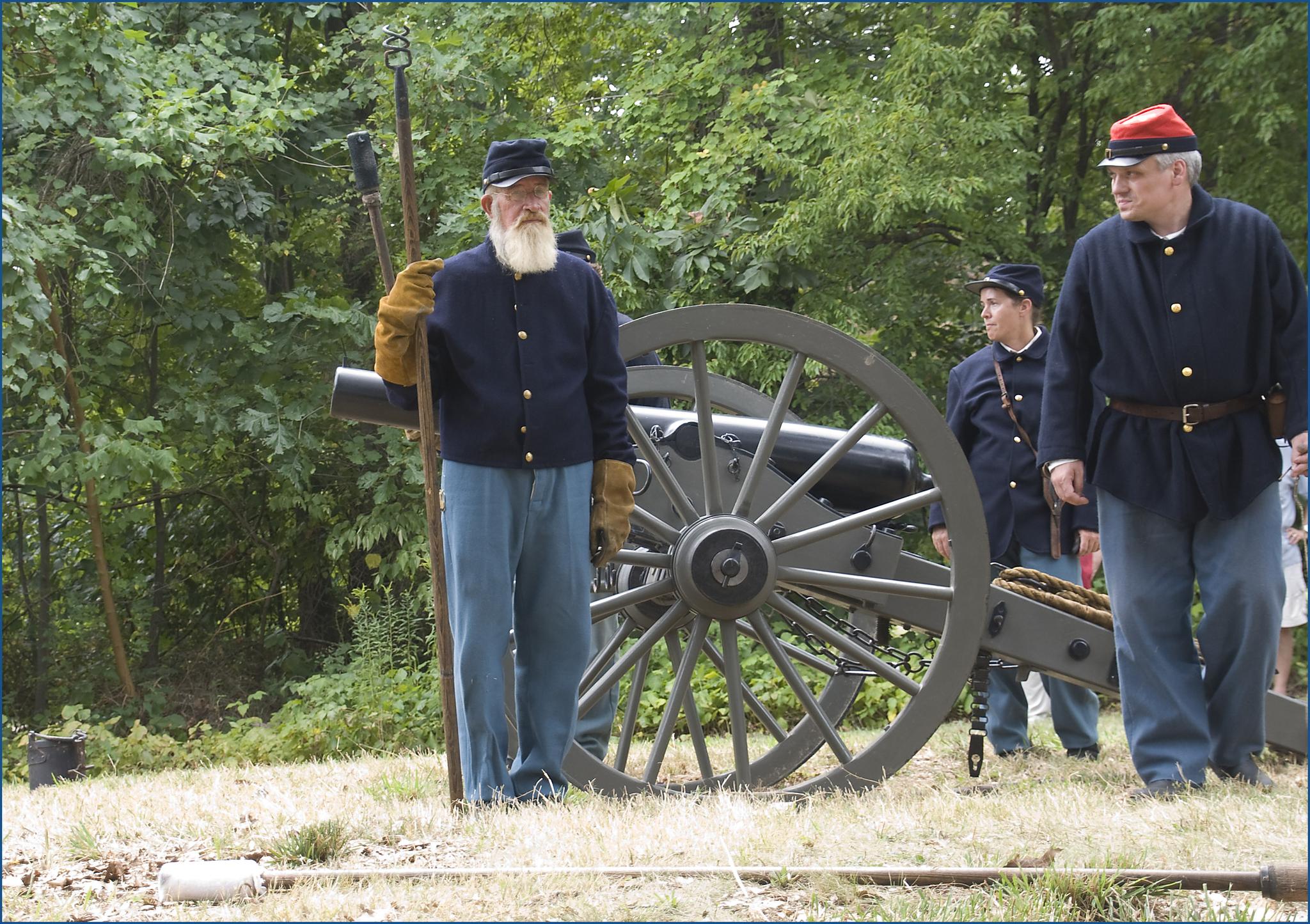 Three men in historical uniforms standing near a large cannon outdoors.