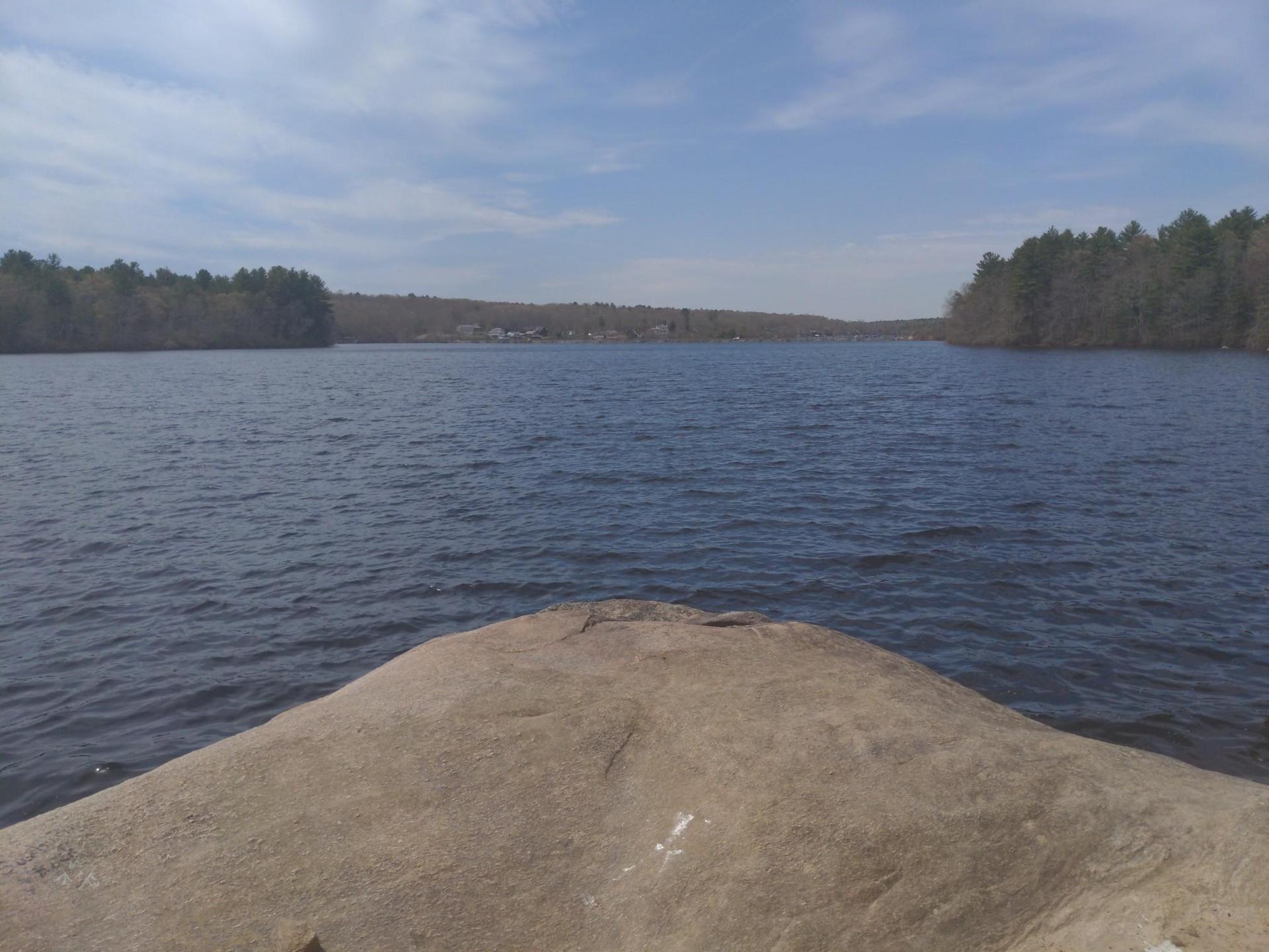 Wide lake view from a rock, surrounded by distant trees under a cloudy sky.
