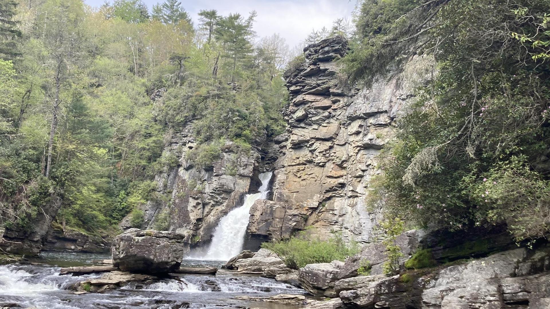 Waterfall cascading over rocks into a river, surrounded by green trees and cliffs.