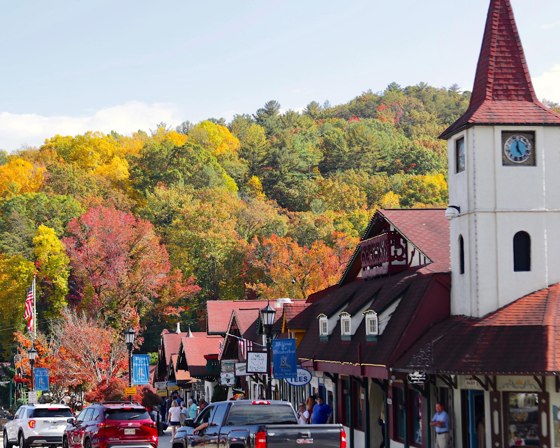 Busy street with shops, cars, and colorful autumn trees on hills.