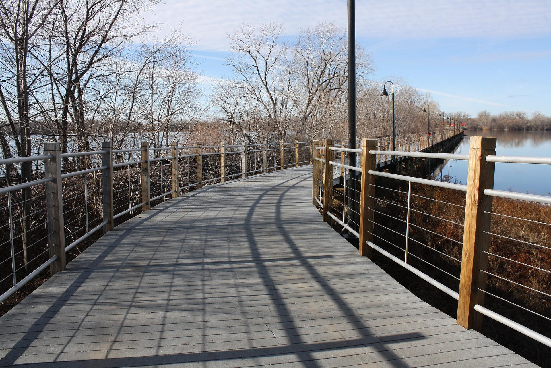 A sunny wooden boardwalk curves over water with bare trees.