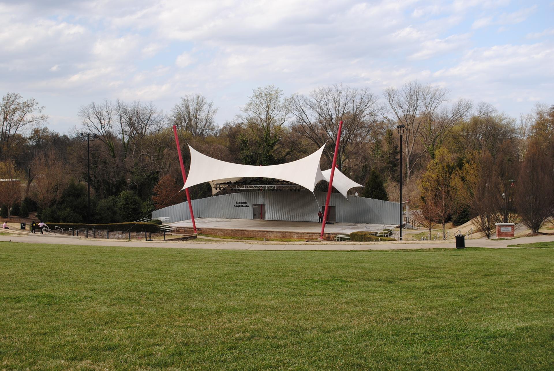 Outdoor amphitheater with white tensile roof, green field, and bare trees under overcast sky.