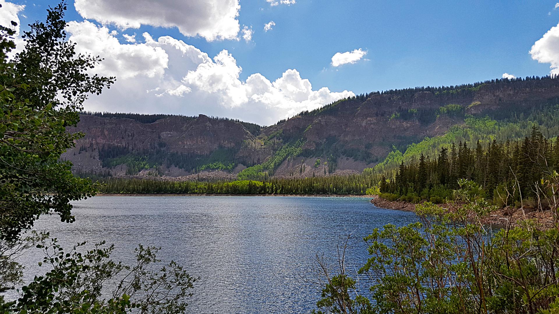 A serene lake surrounded by forested mountains under a bright, partly cloudy sky.