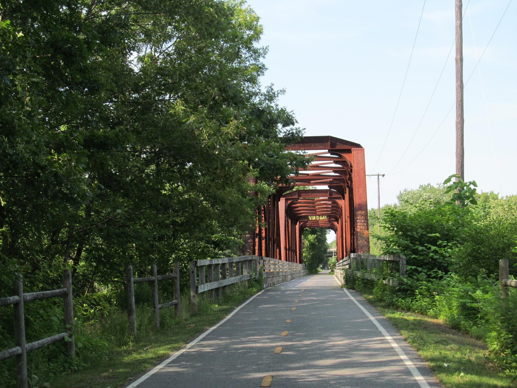 Paved path leading to a red metal truss bridge on a sunny day.