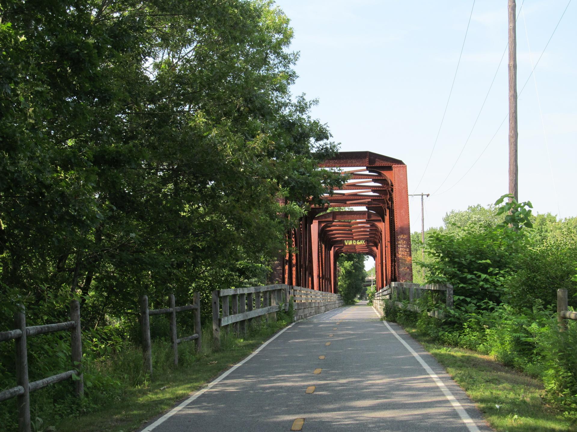 Paved path leading to a red metal truss bridge on a sunny day.
