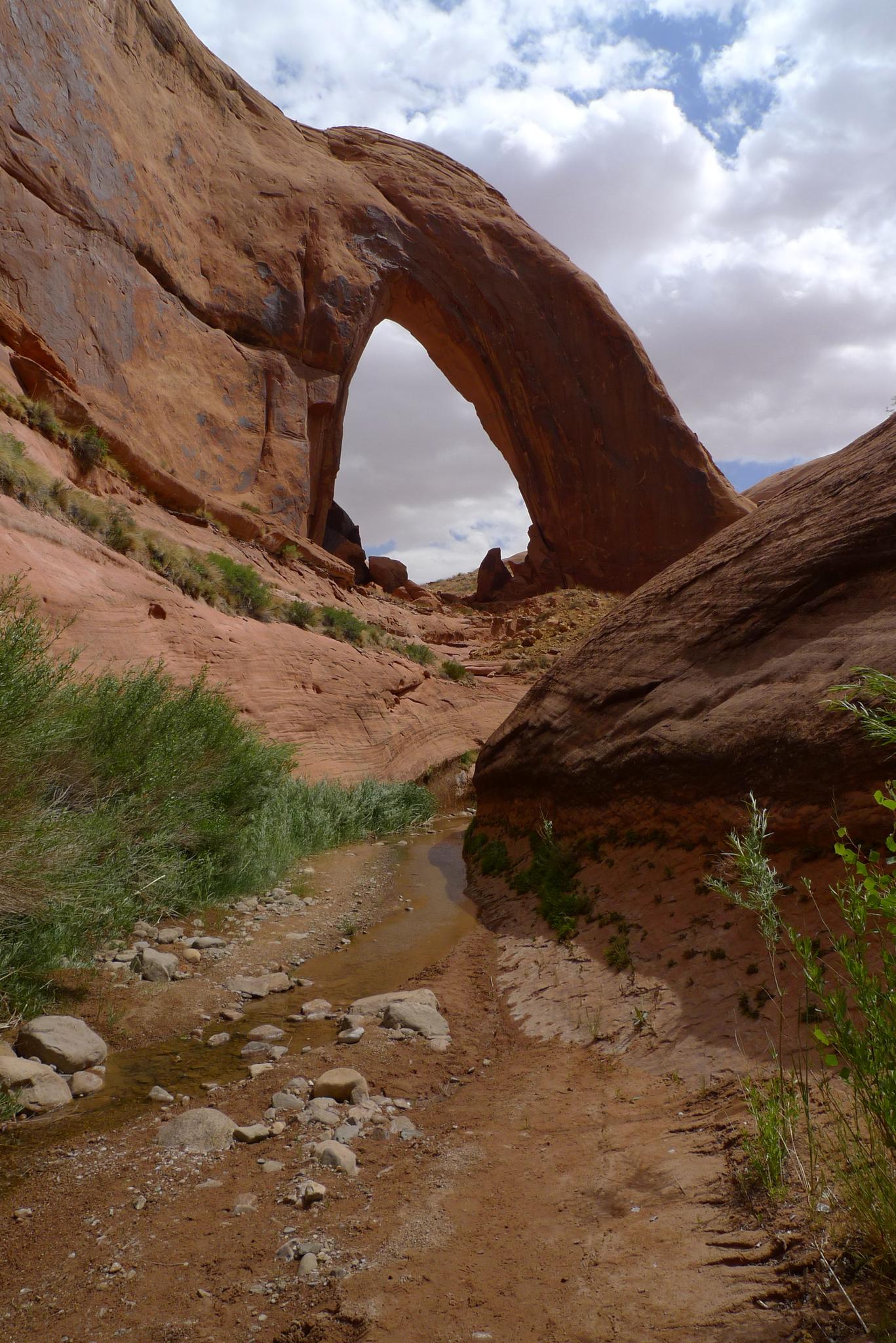A natural rock arch spans over a stream and rocky path under cloudy skies.