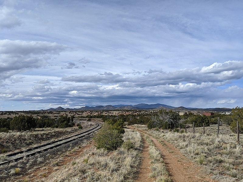 Railroad tracks and a dirt road wind through arid scrubland under a cloudy sky.
