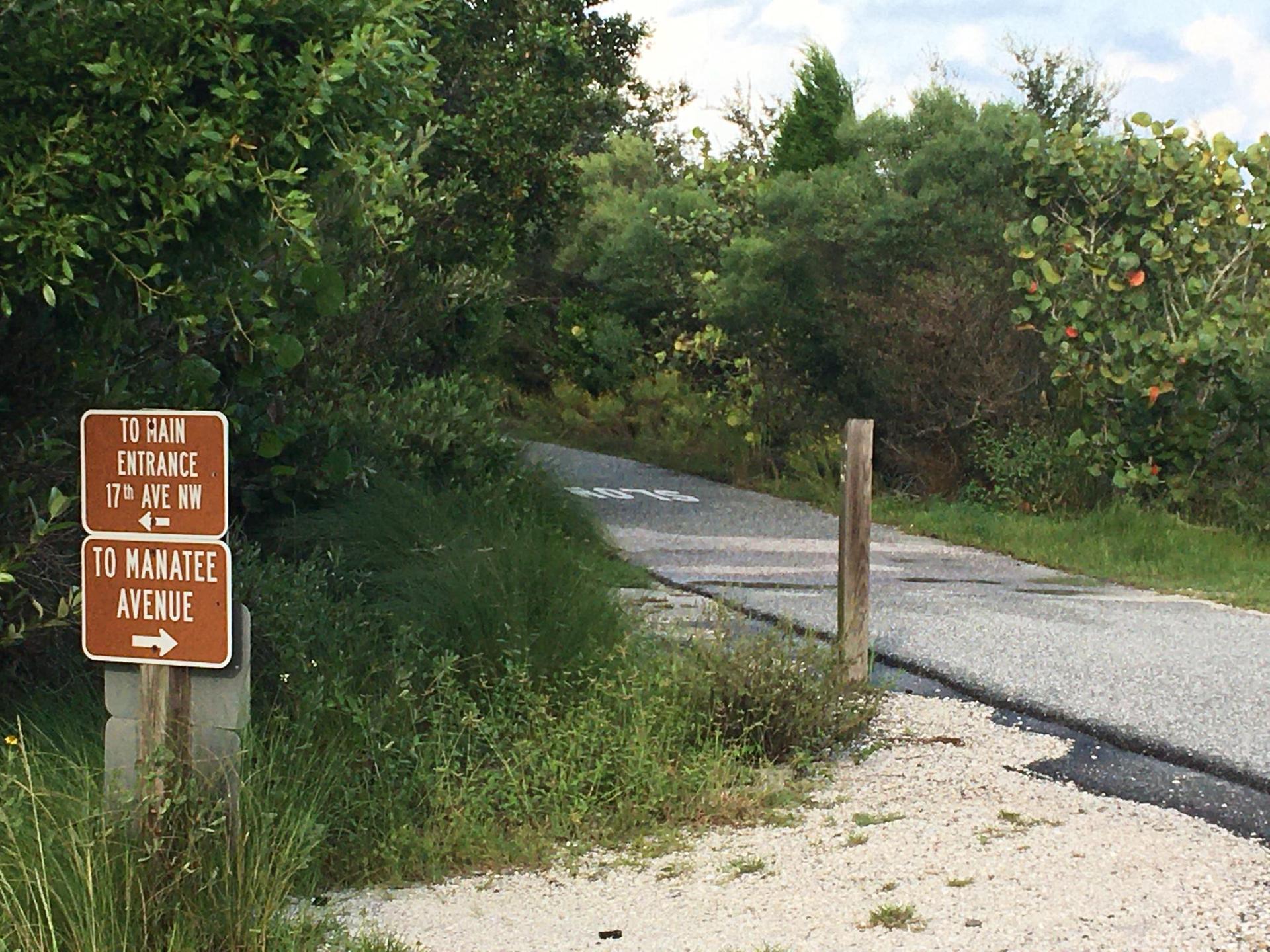 A brown directional sign on a gravel path next to an asphalt road.