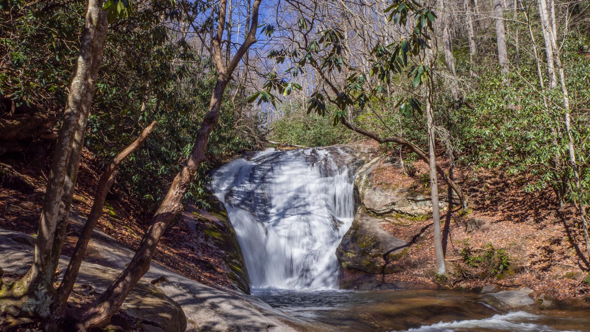 Waterfall cascading into a rocky stream, surrounded by sunlit trees.