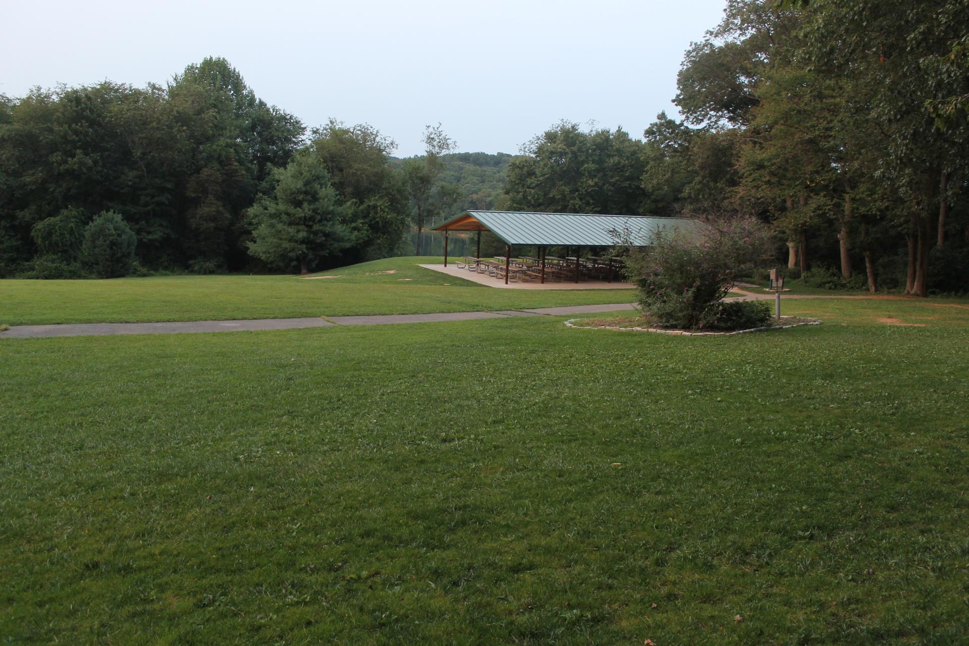 Large grassy field with a covered pavilion and distant trees under an overcast sky.