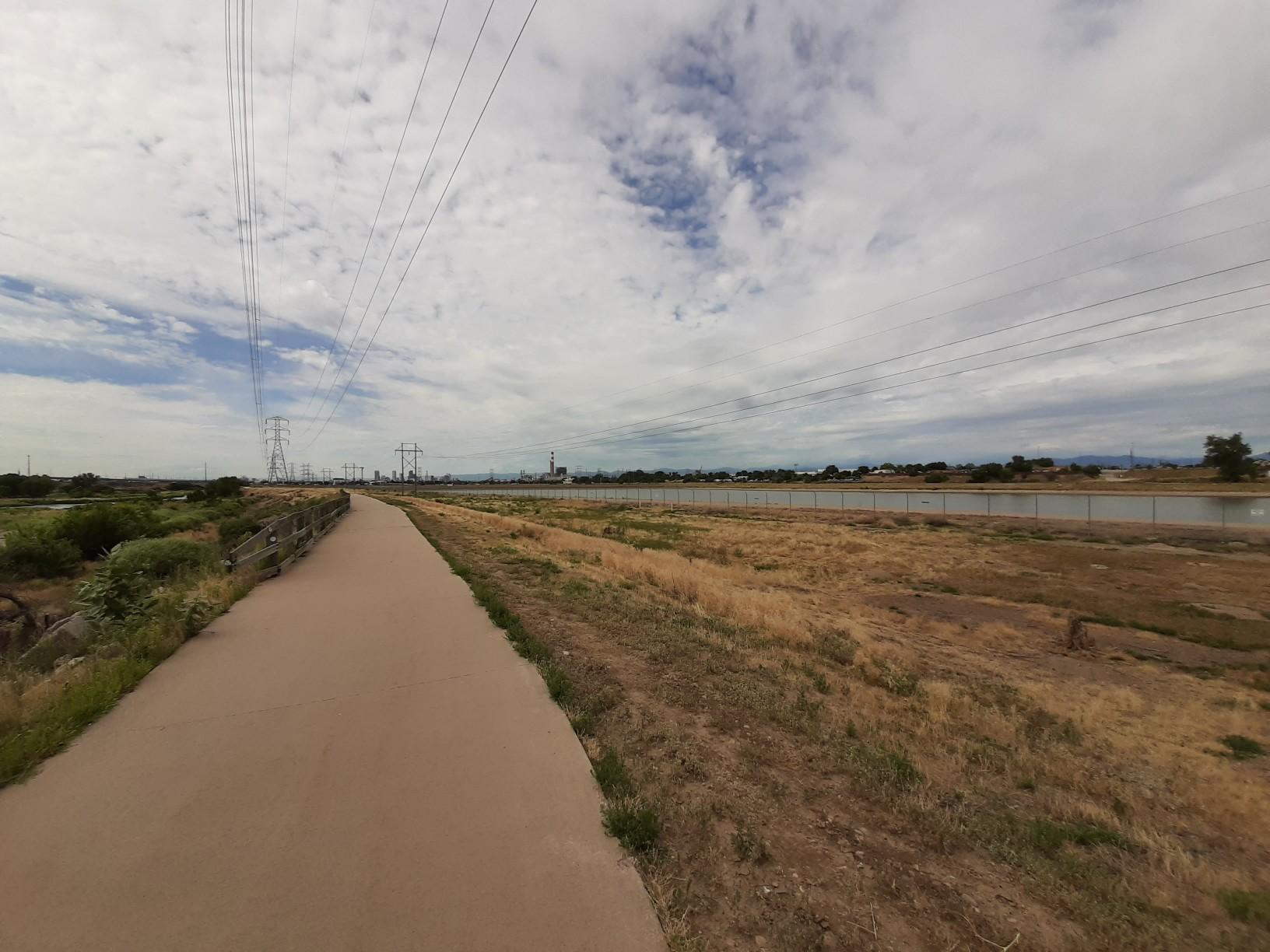 A paved path runs alongside a dry field and water under a cloudy sky.