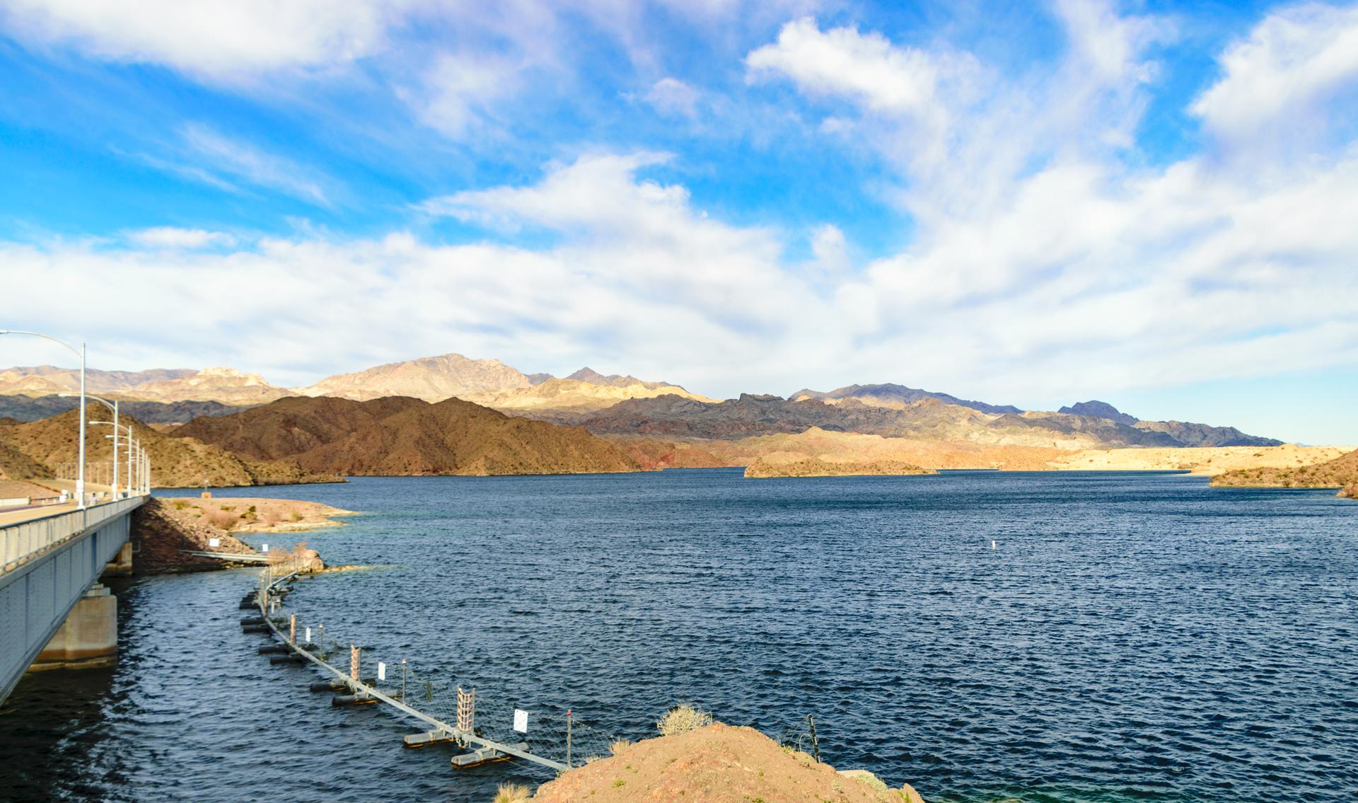 Expansive blue lake with a bridge, rugged mountains, and a cloudy sky.