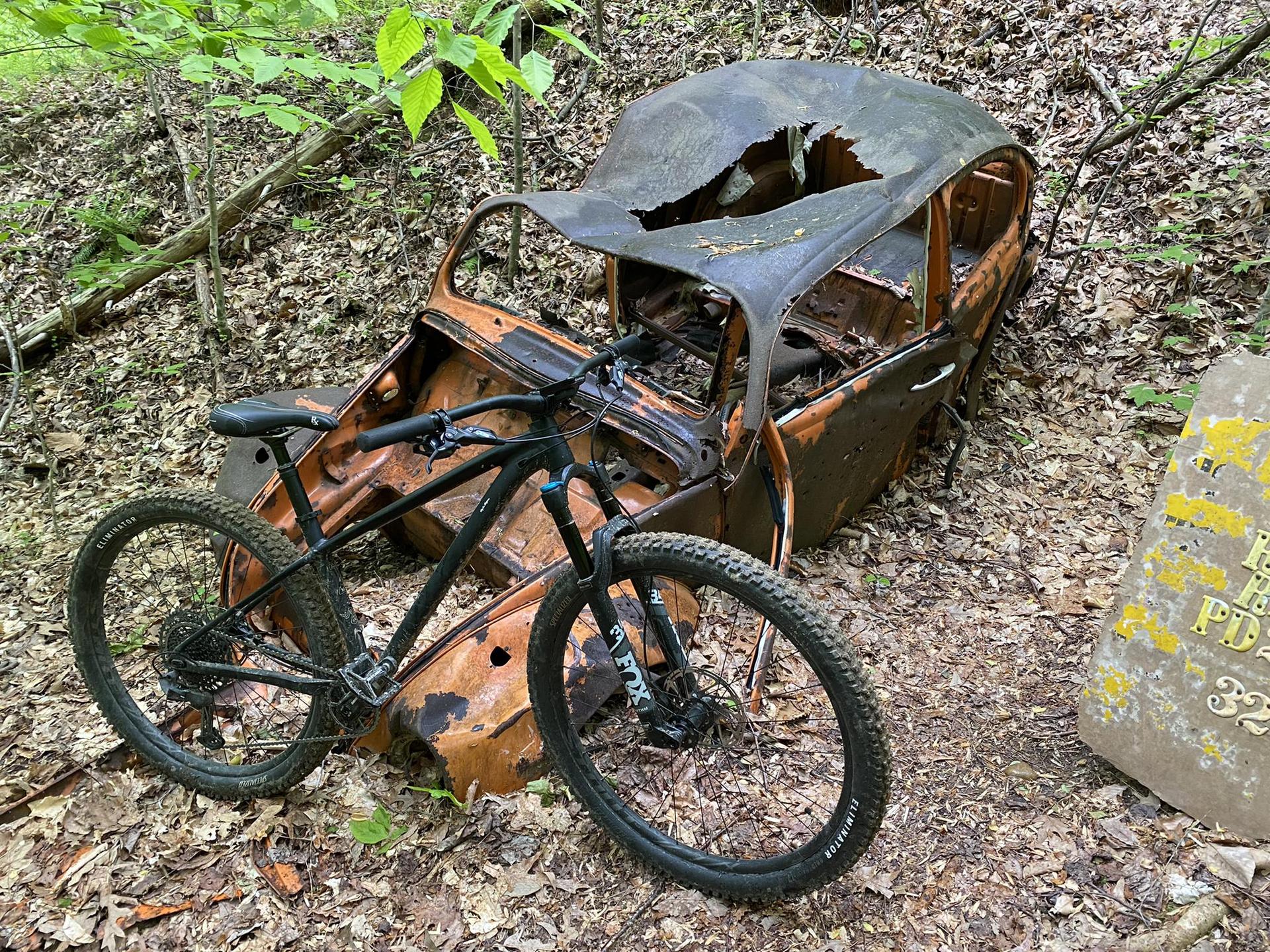 A black mountain bike rests next to a rusty, abandoned car in the woods.