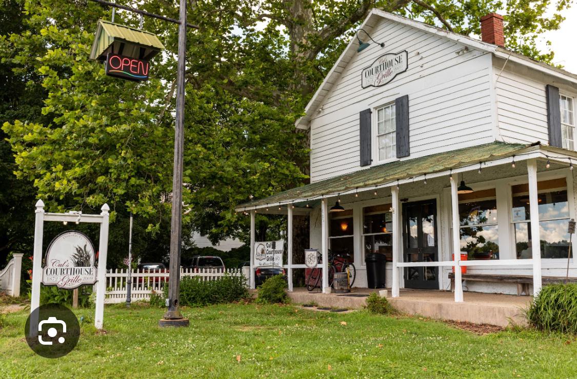 White clapboard building with a porch, an 'Open' sign, and green lawn.