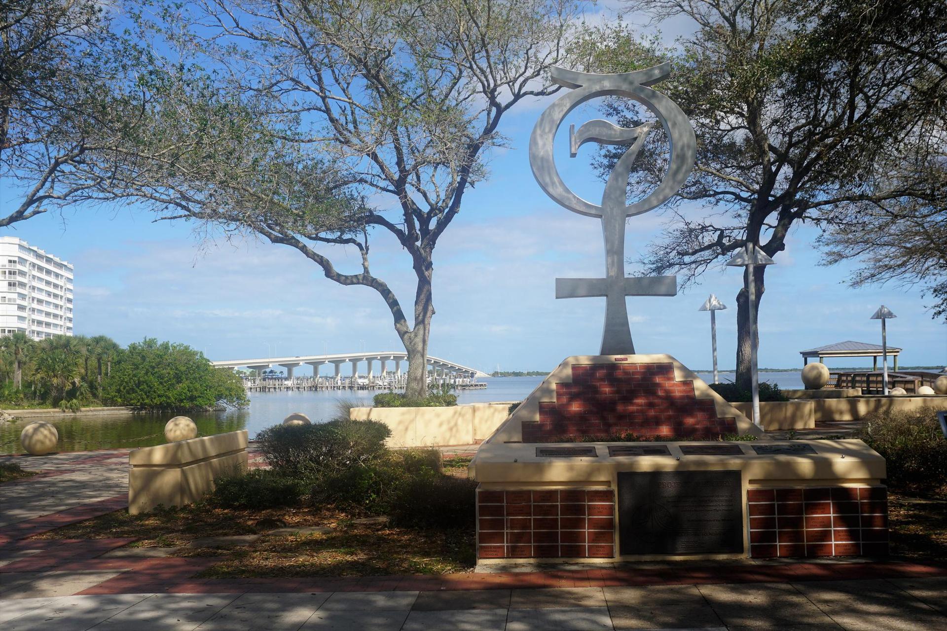 Large outdoor sculpture near water, with trees and a distant bridge.