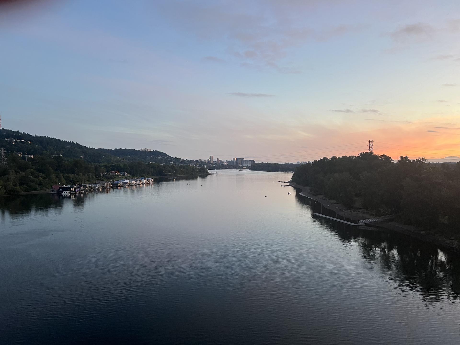 Wide river at sunset with forested banks and distant city skyline.