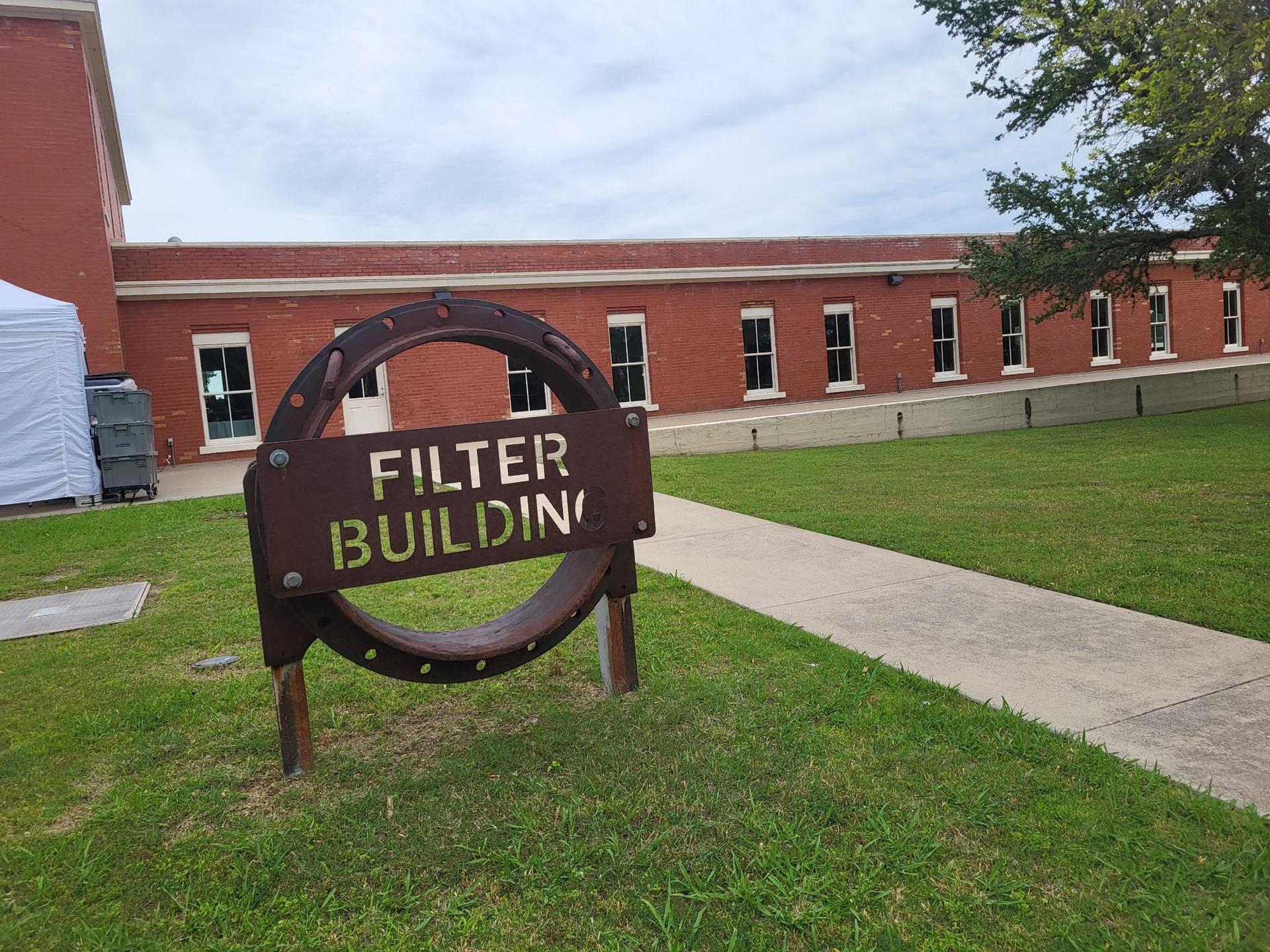 A brown sign on a green lawn in front of a long red brick building.