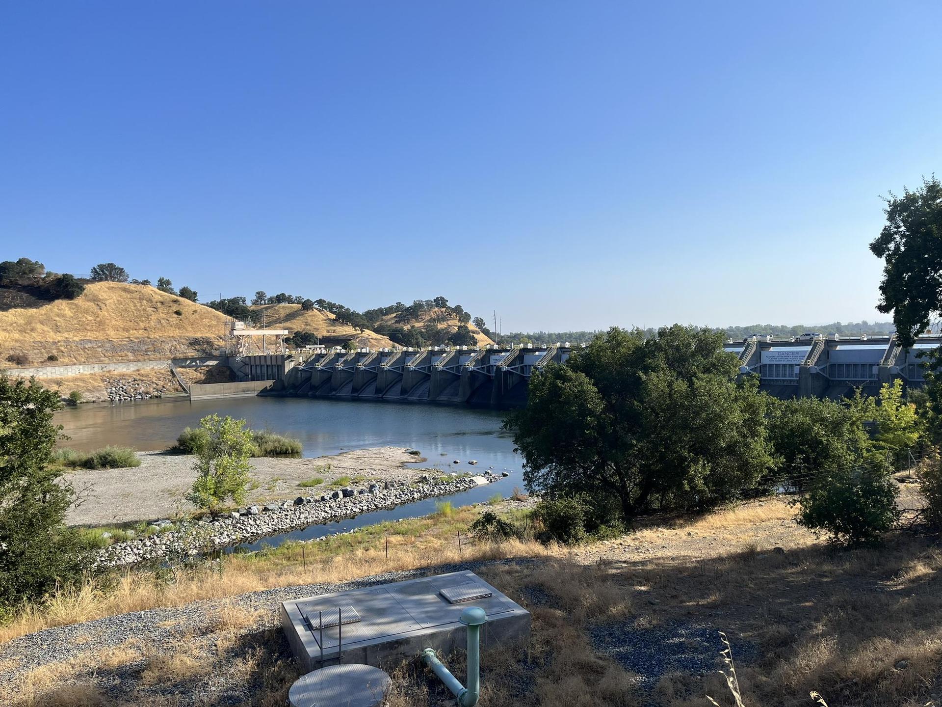A large concrete dam and river in a hilly landscape under a clear blue sky.