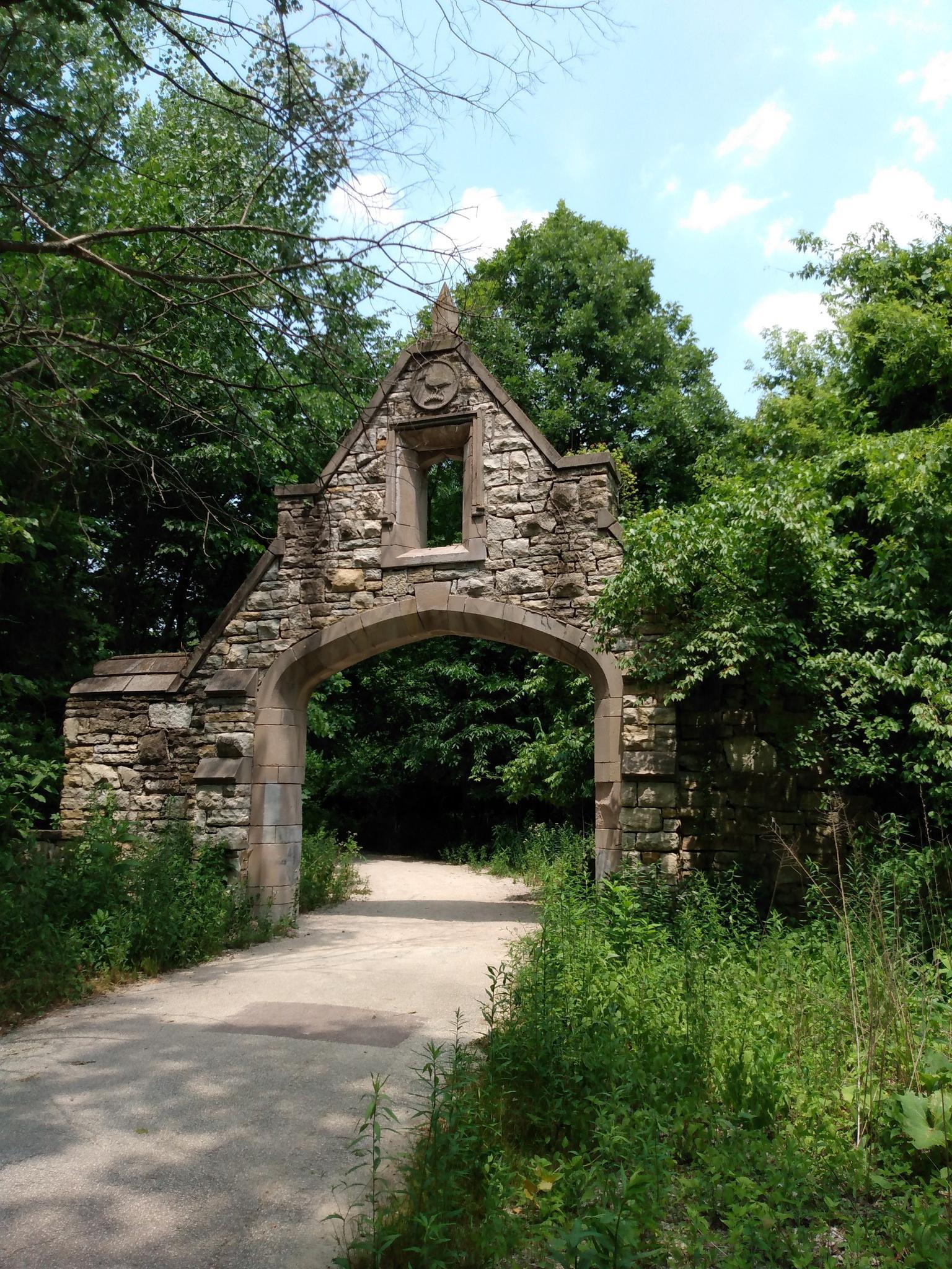 Ornate stone archway over a dirt path, surrounded by lush green trees.