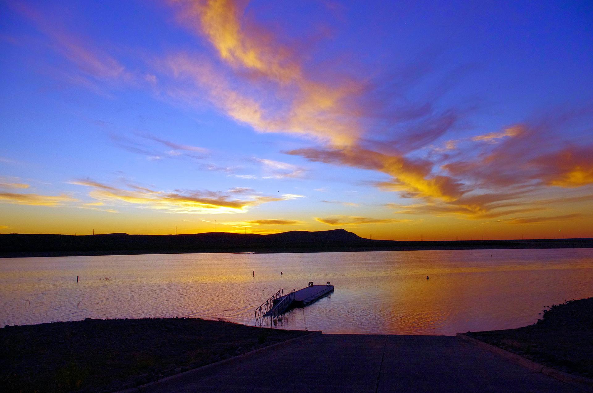 Vibrant sunset over a calm lake with a boat dock and distant hills.