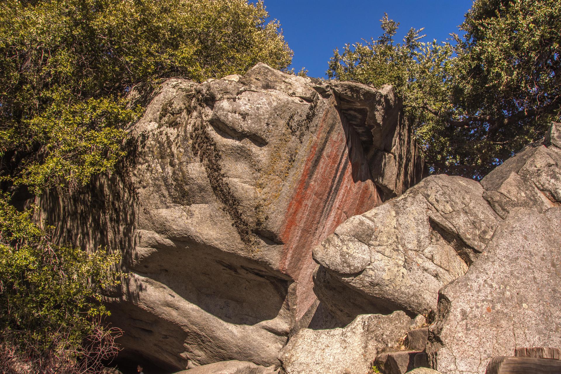 Massive rock formations with striking red streaks, surrounded by green trees.