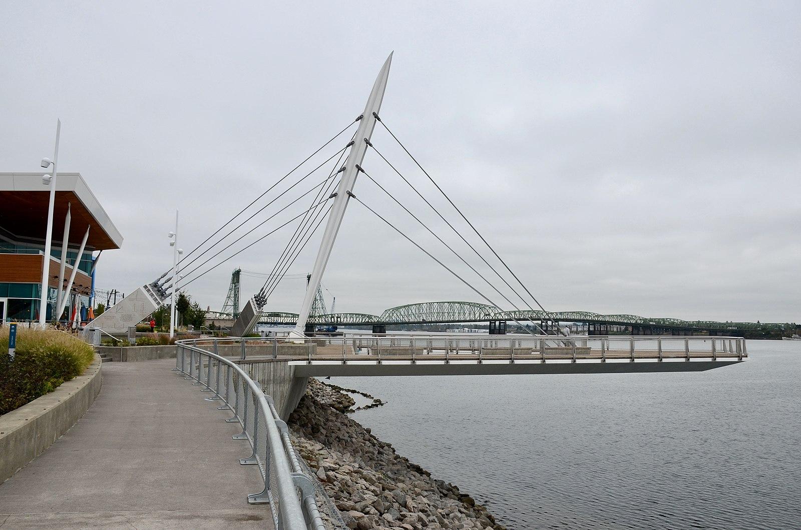 Modern pedestrian bridge over water, with distant bridges, on an overcast day.