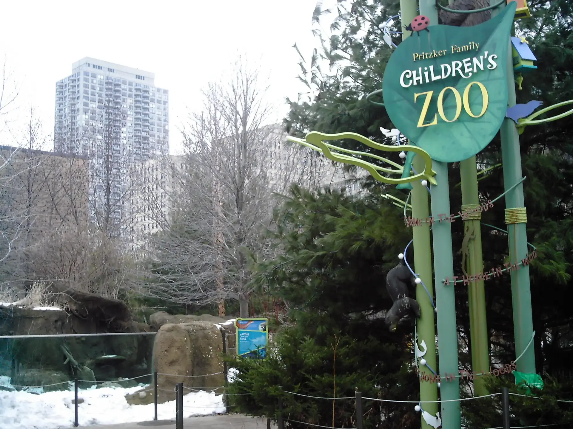 Children's zoo sign in winter with snow, bare trees, and a tall building.