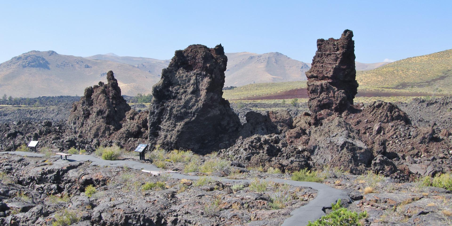 Boardwalk path winds past large volcanic rock pillars on a sunny day.