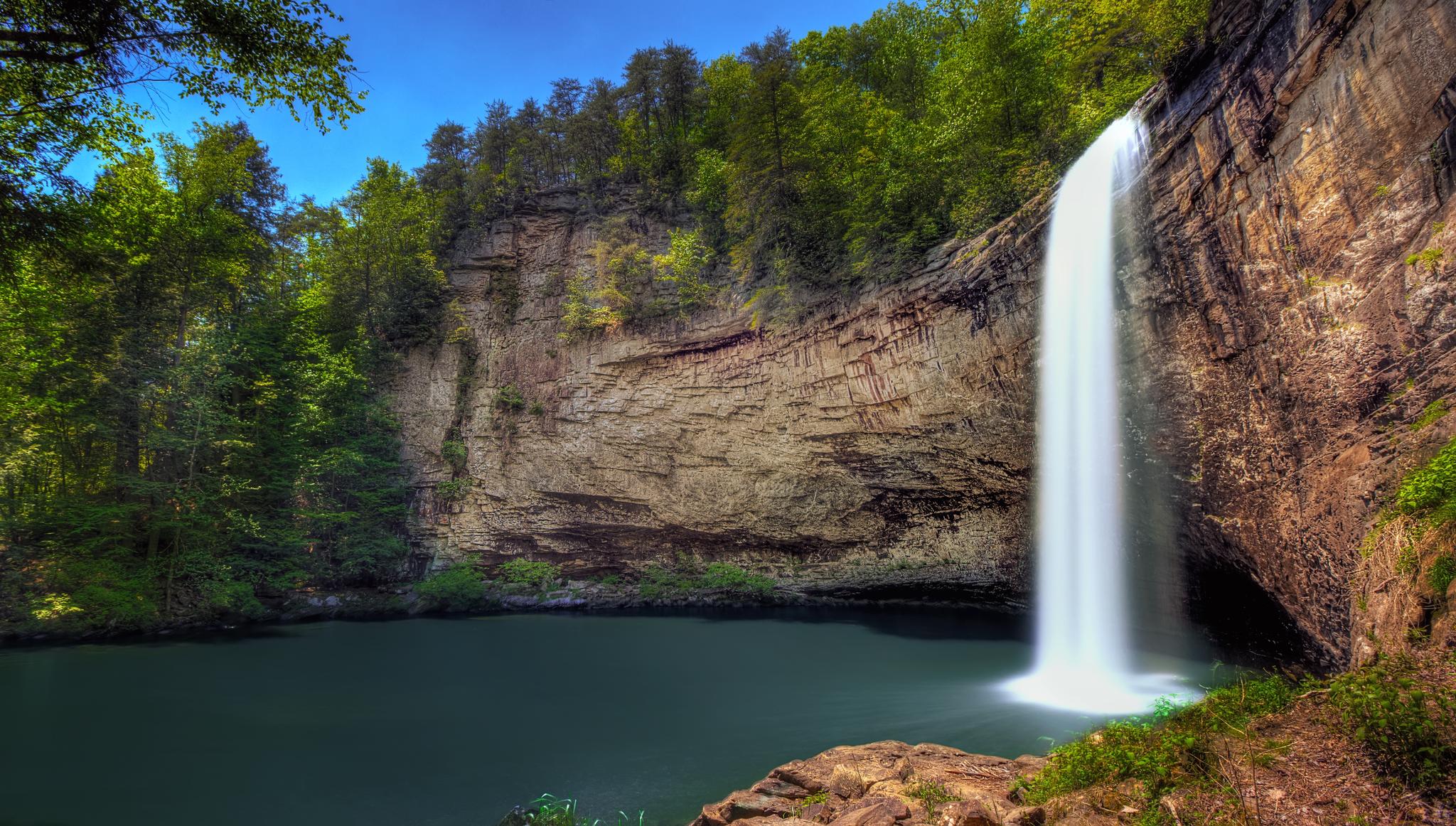 A powerful waterfall cascades into a dark pool surrounded by cliffs and green trees.