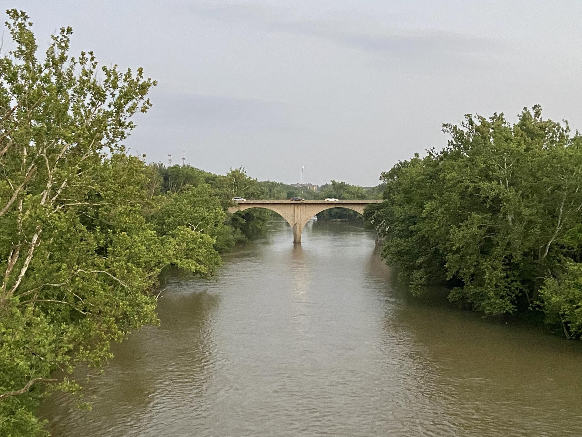 Wide river flowing under a distant arched bridge, flanked by green trees.