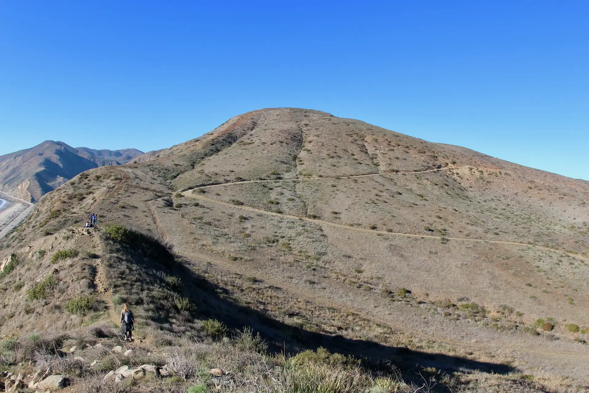 Hikers ascend a winding trail on arid, sunlit hills under a clear blue sky.