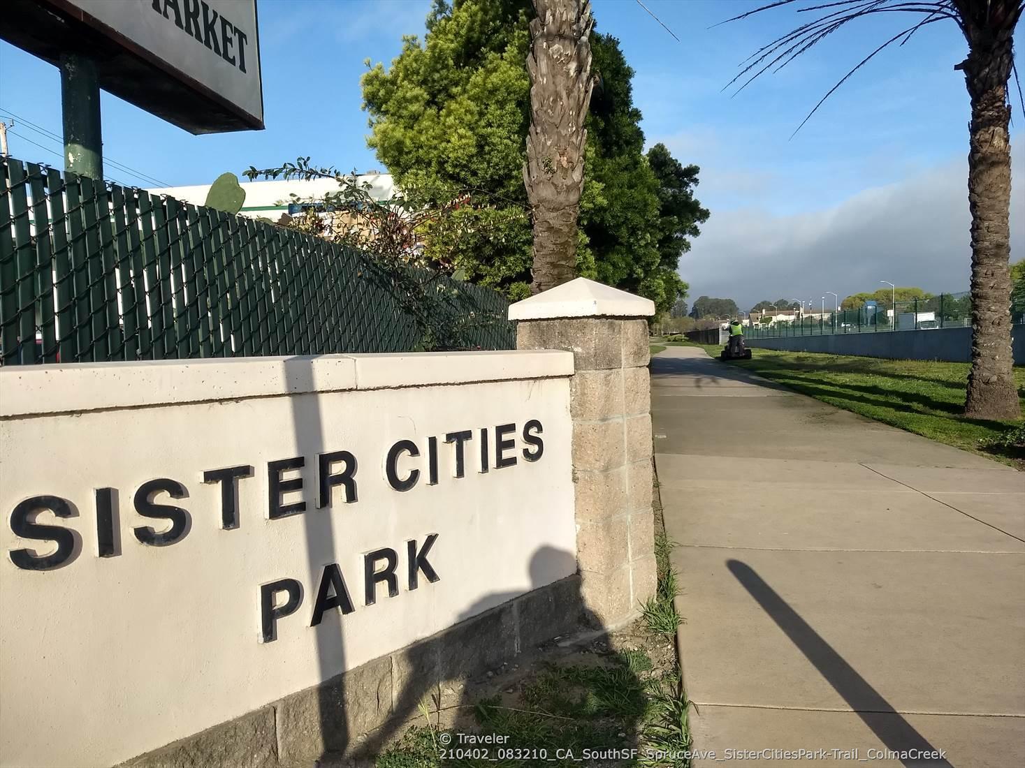 A stone sign reads "Sister Cities Park" next to a sidewalk and palm trees.