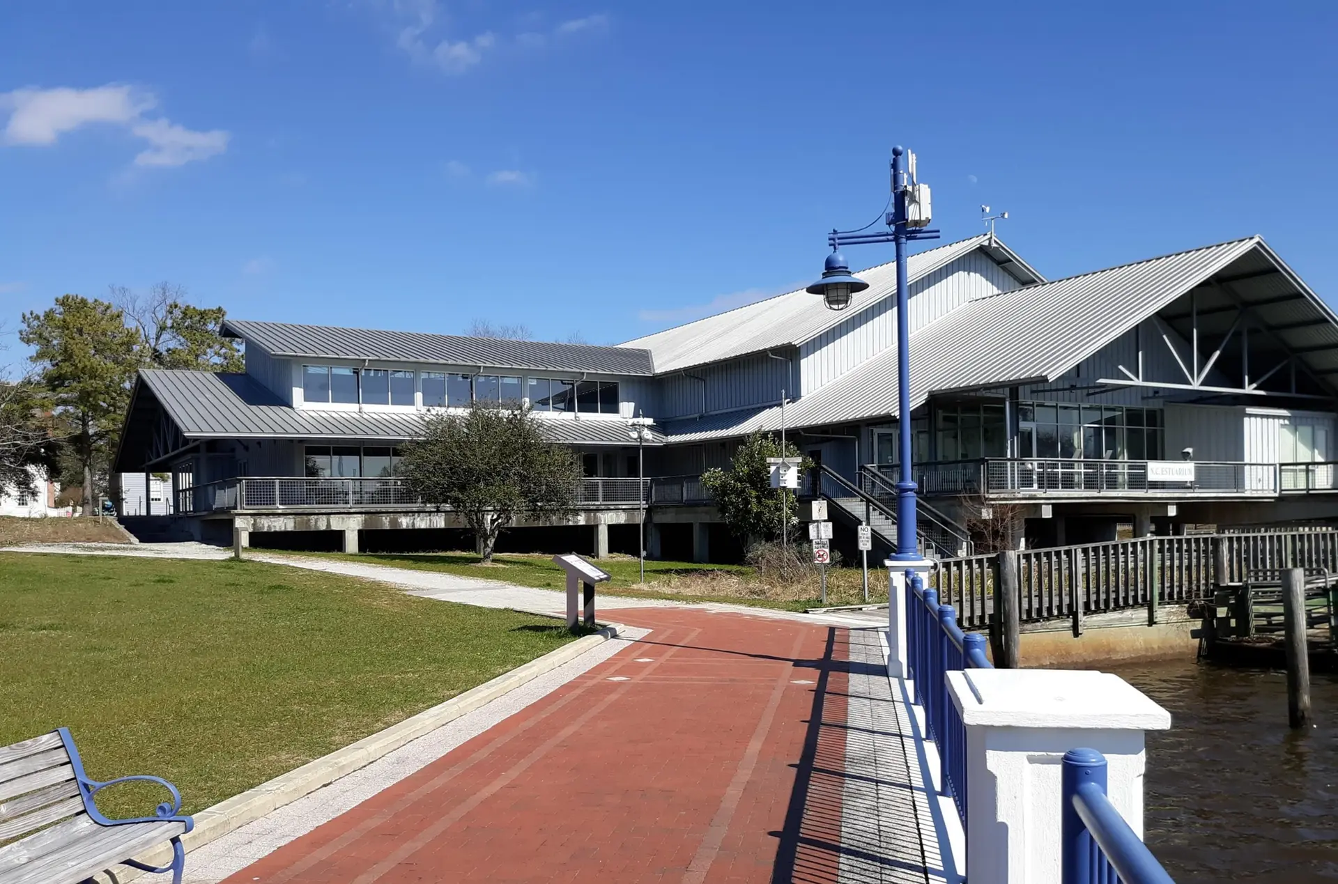 Large building with metal roof next to a paved path and water on a sunny day.