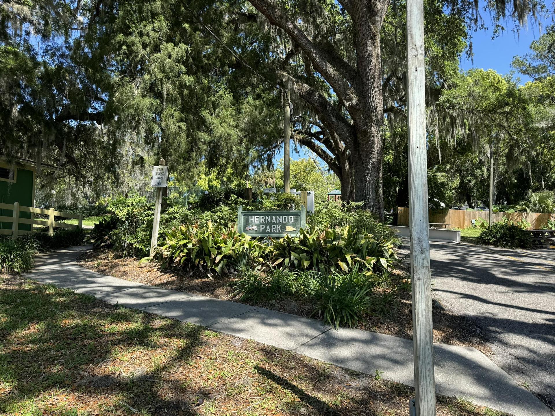 Sunny view of a park entrance sign surrounded by green foliage and large trees.