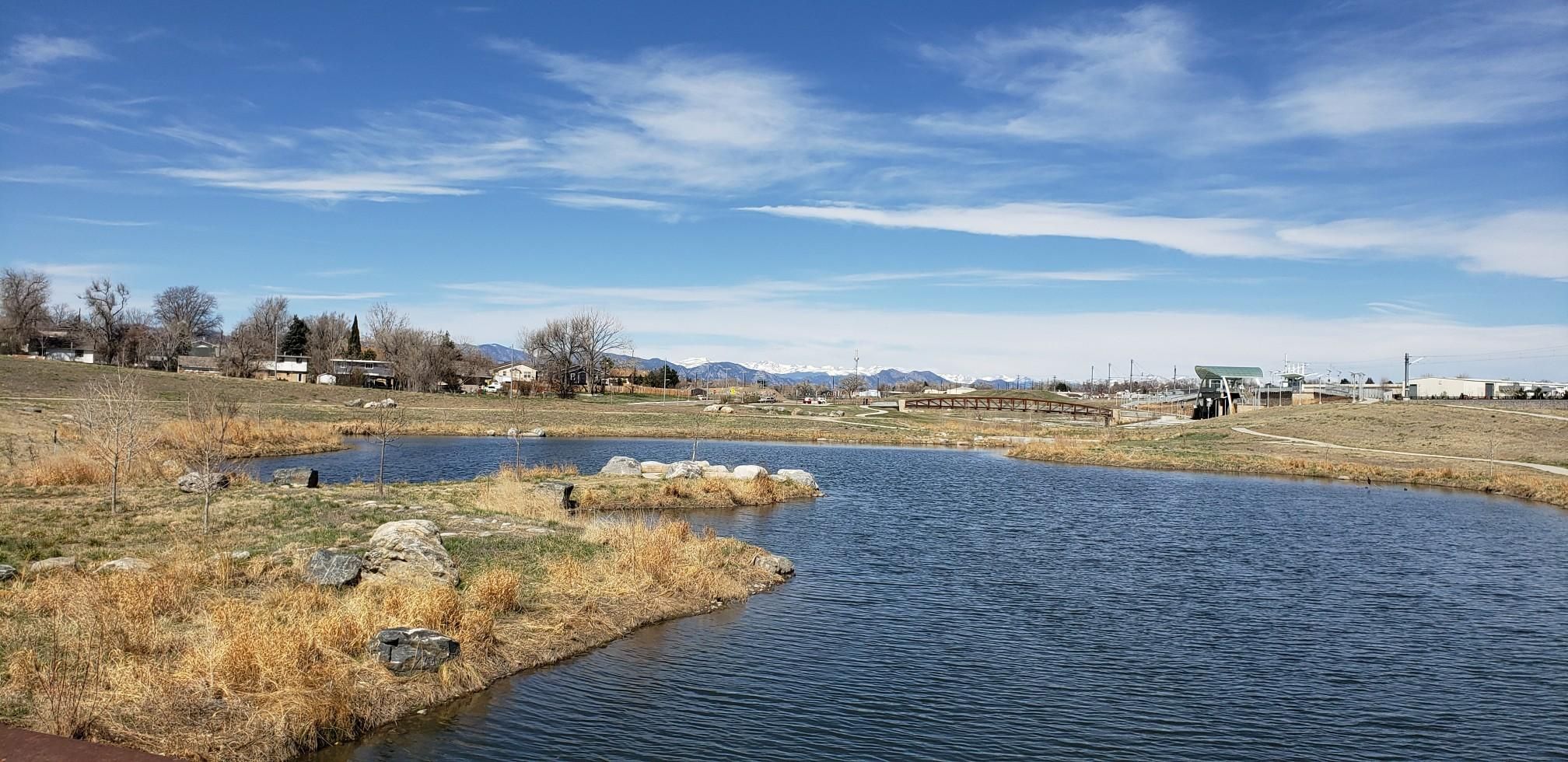 A pond with dry grassy banks under a bright blue sky with distant mountains. - Big Dry Creek – Westminster Station Pond loop from Westminster