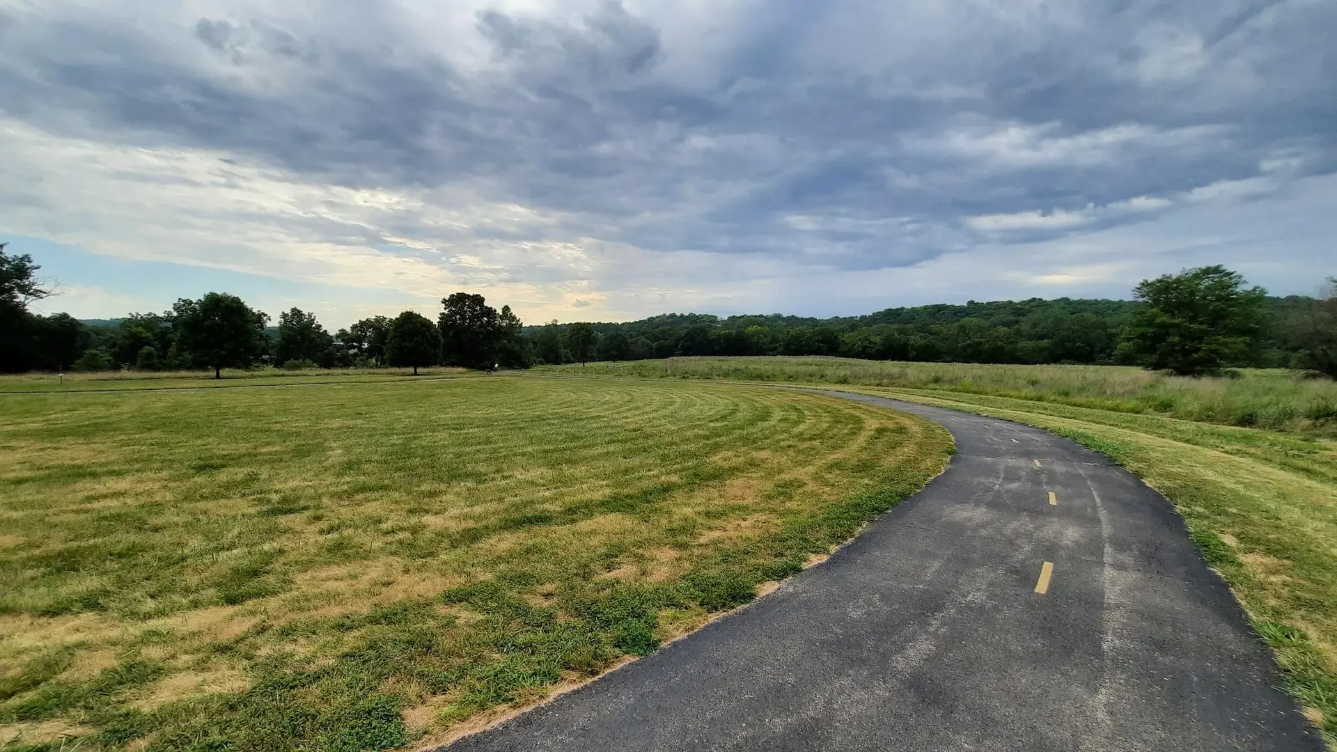 Paved path curves through a large green field under a cloudy sky.