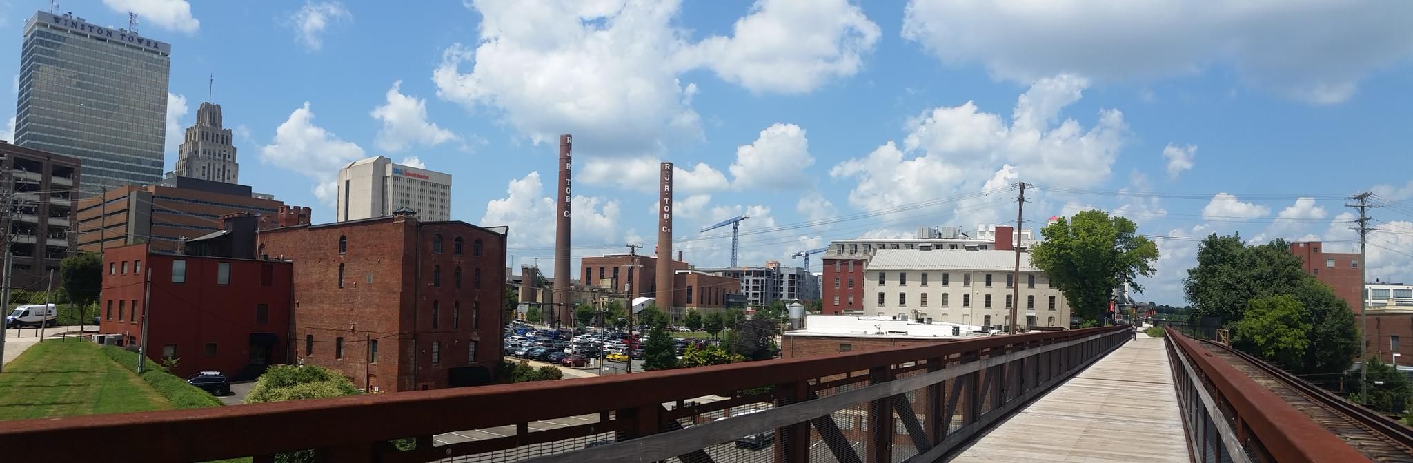 Pedestrian bridge overlooking a city skyline with brick buildings under a sunny sky. - Long Branch Trail – Reynolda Gardens loop from Country Club Road & Knollwood Street