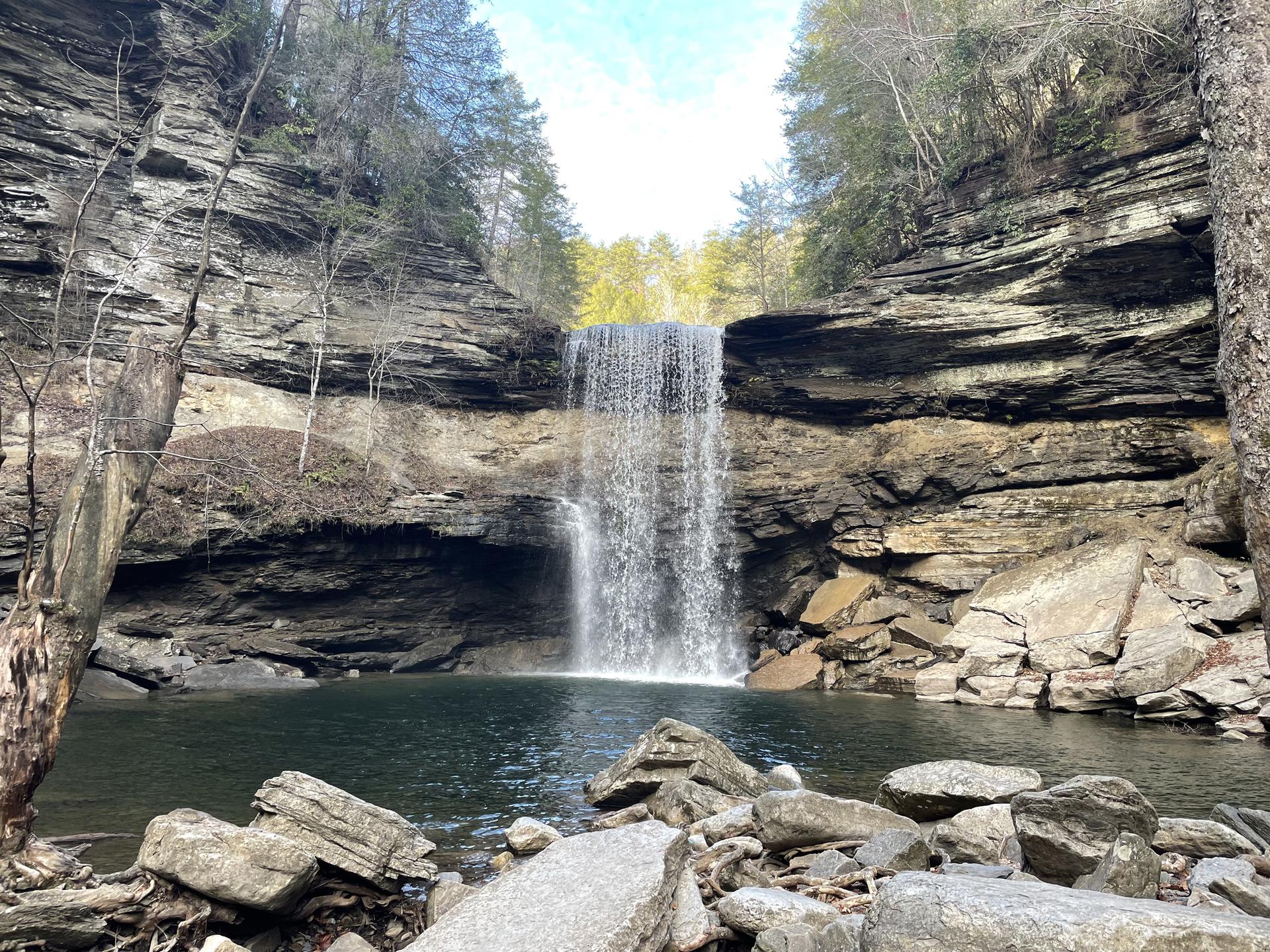 A wide waterfall plunges into a dark pool surrounded by rocky cliffs and trees.