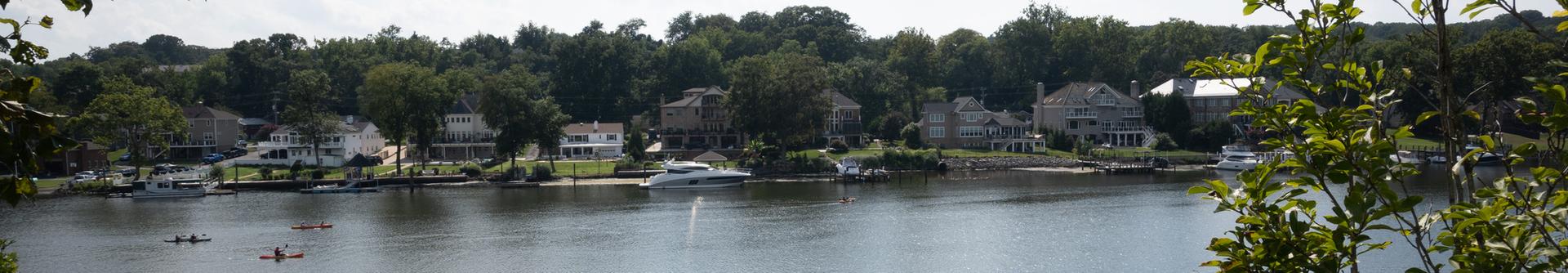 Wide river with houses, boats, and kayakers under bright daytime light.