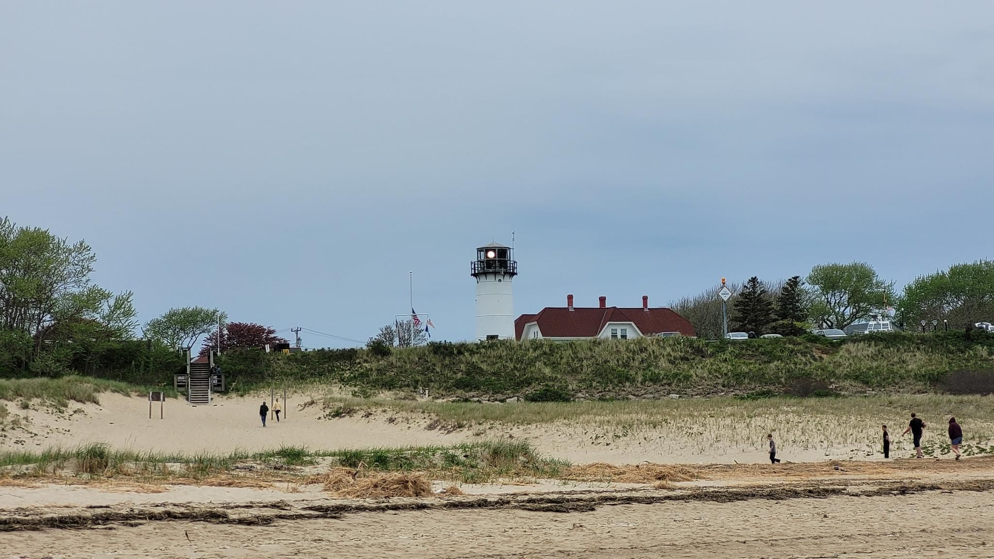 People on a sandy beach with a distant lighthouse under an overcast sky. - Lighthouse Beach Trail