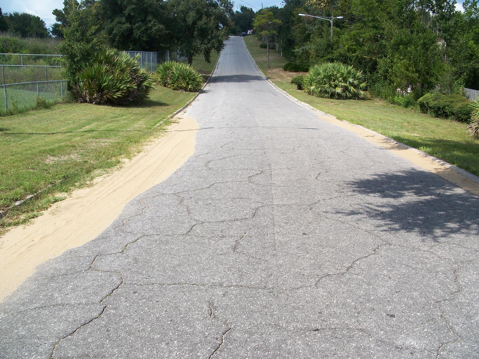 Cracked asphalt road going uphill, flanked by grass, sand, and trees.