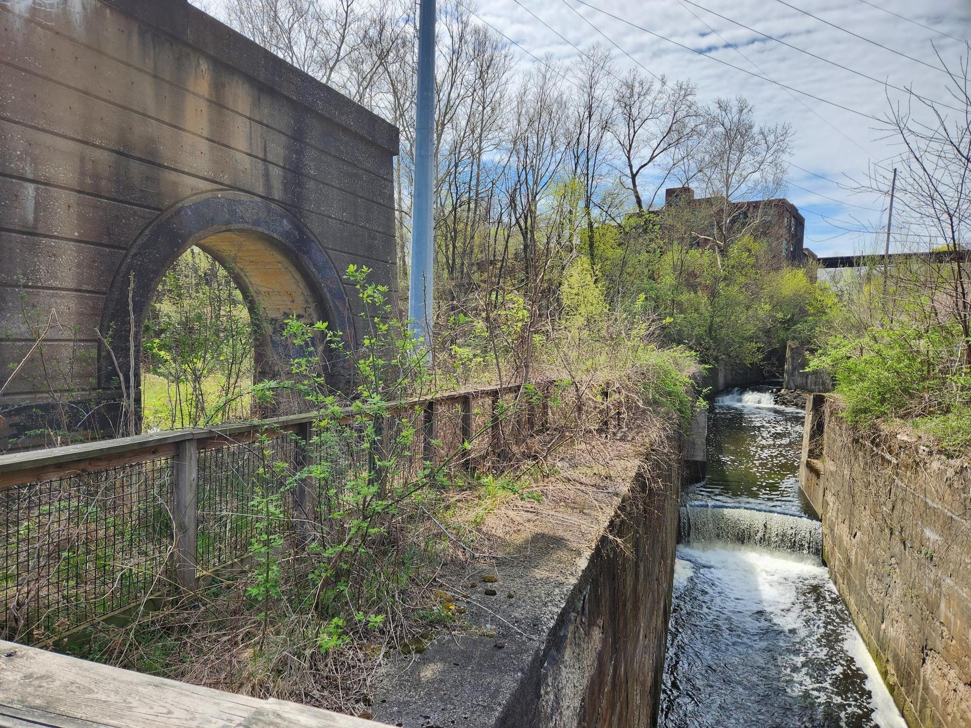 Stone archway and concrete structures next to a flowing stream, under a cloudy sky.