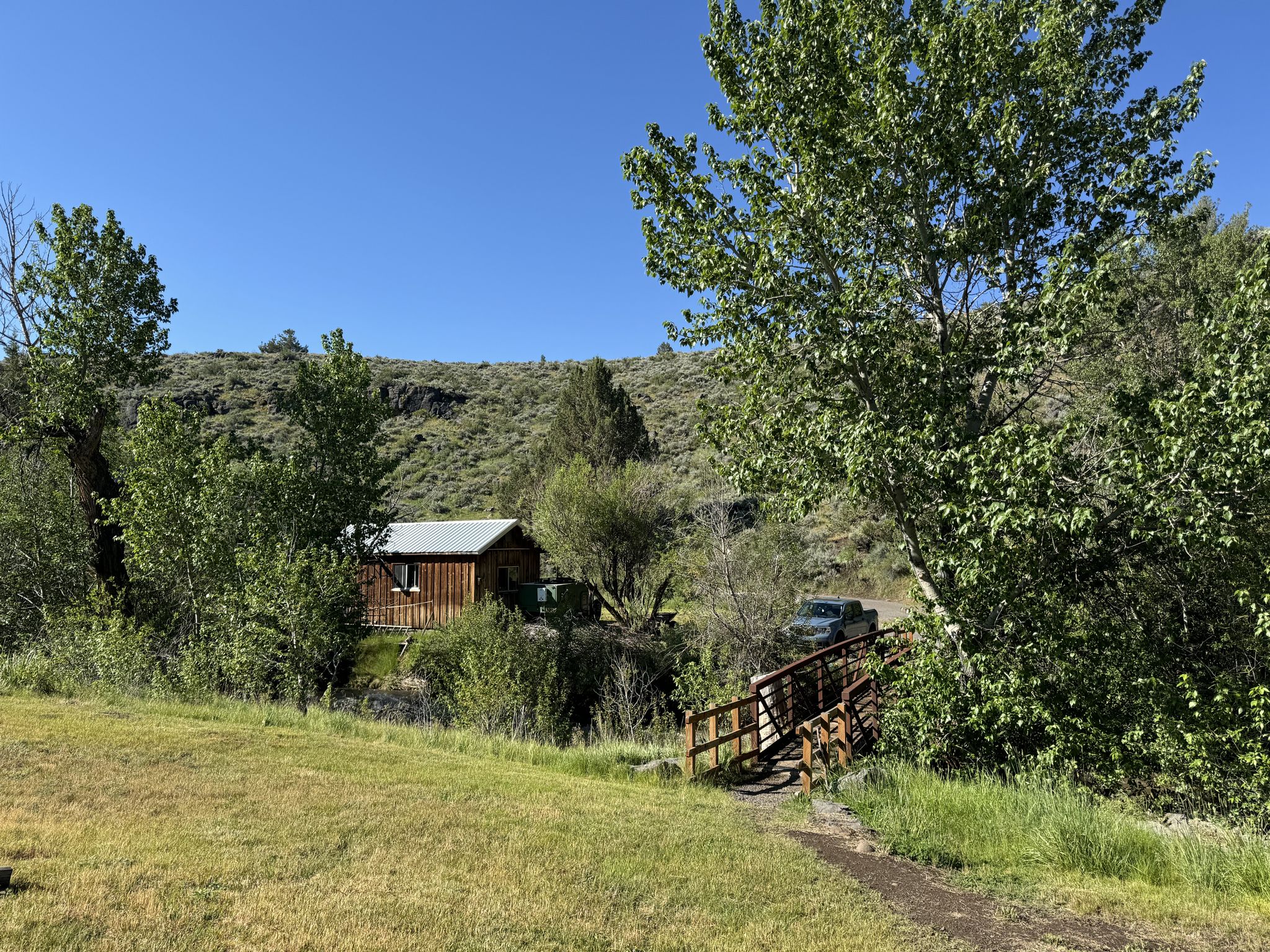 Small wooden cabin nestled among trees, with a bridge, under bright sun. - Levi Brinkley Trail