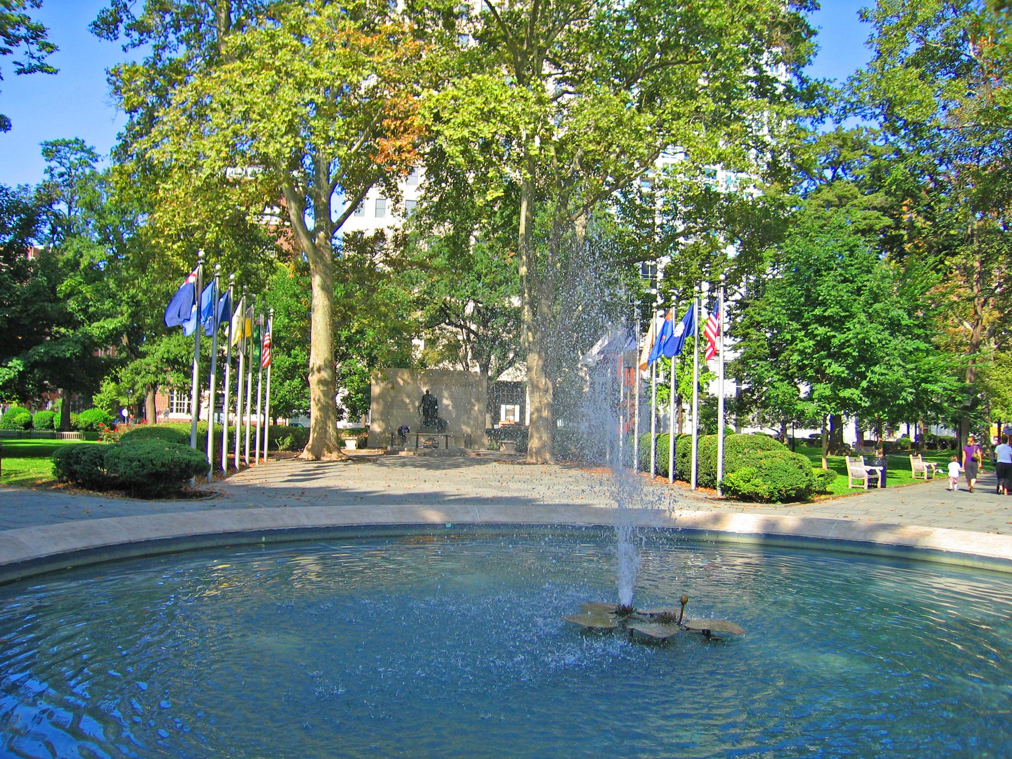 Sunny park scene with a circular fountain spraying water and green trees.