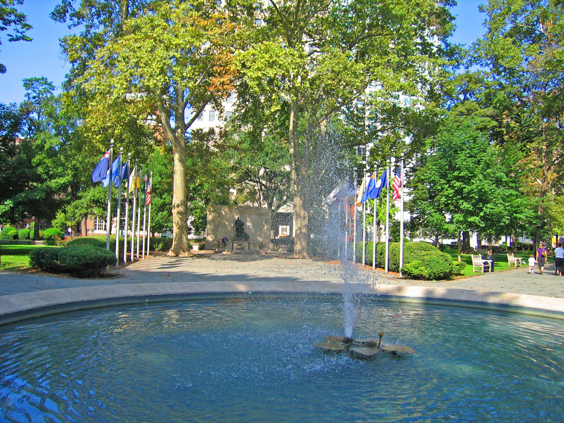 Sunny park scene with a circular fountain spraying water and green trees.