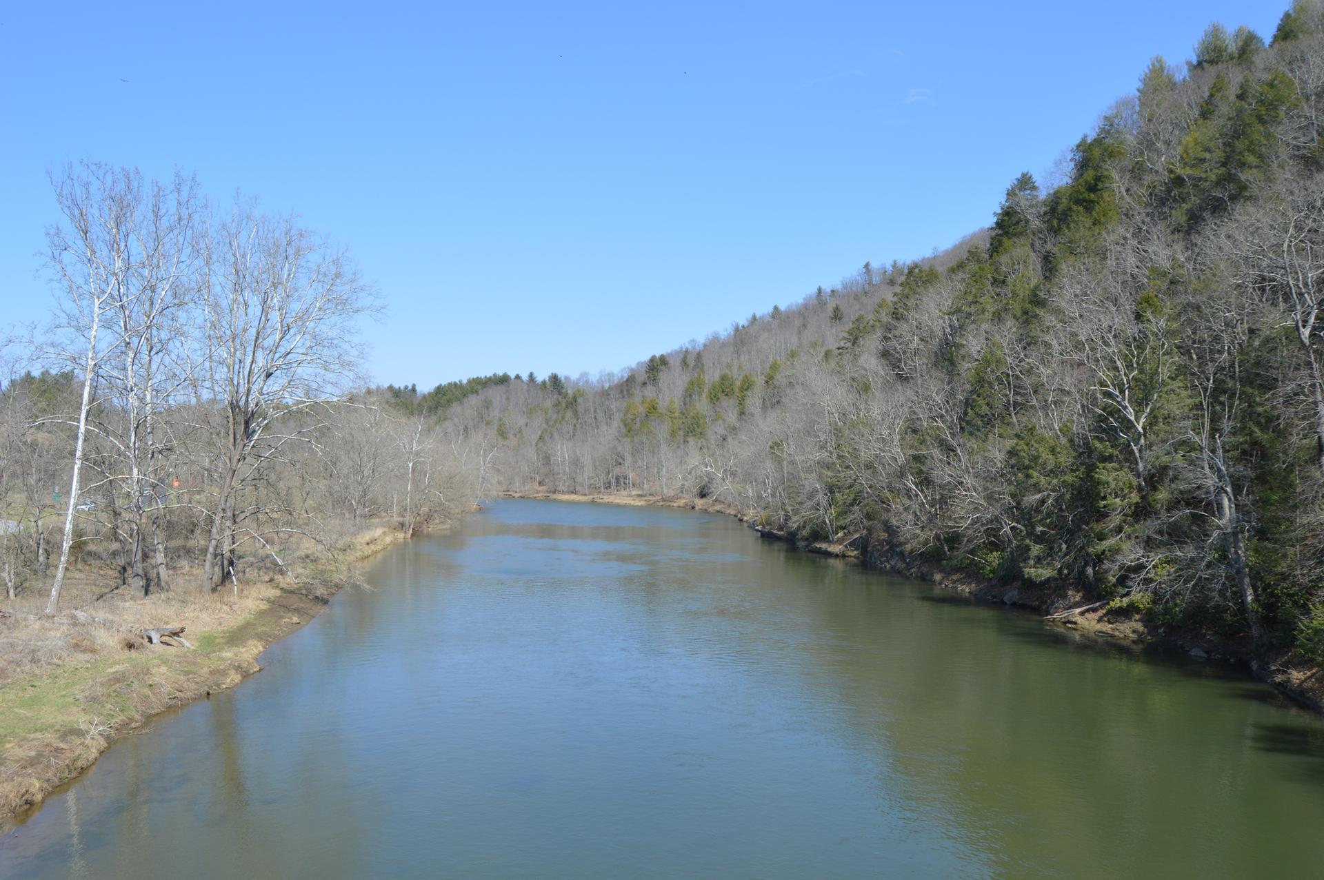 A calm river flows between forested hills with bare trees under a clear sky.