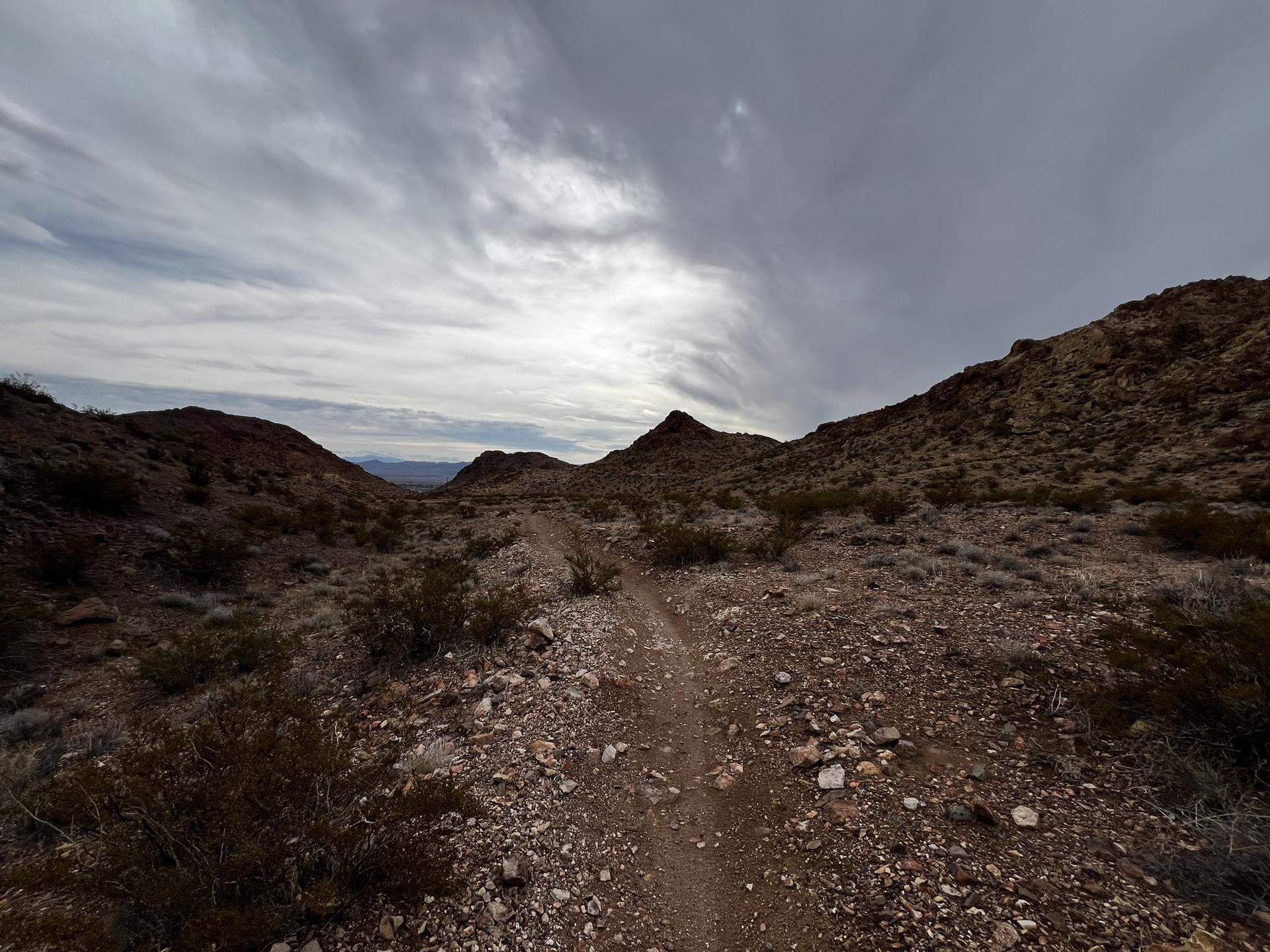 A narrow dirt trail leads through rocky desert mountains under a cloudy sky.