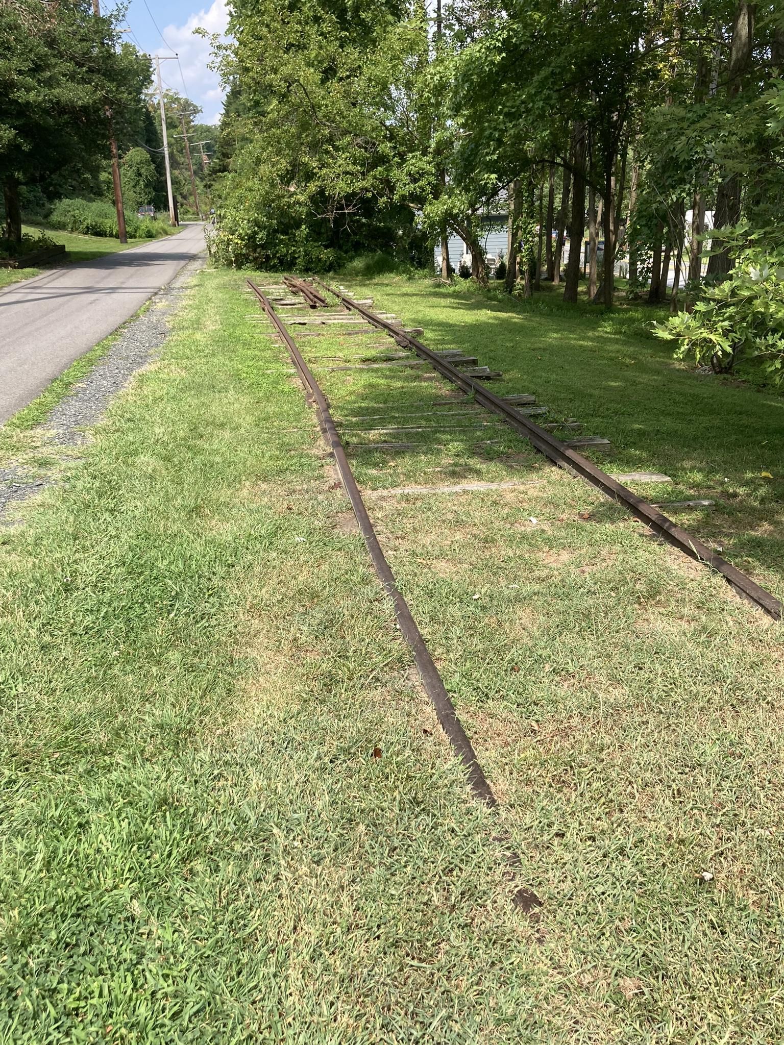 Old train tracks run alongside a paved road in bright daylight. - Baltimore & Annapolis Trail